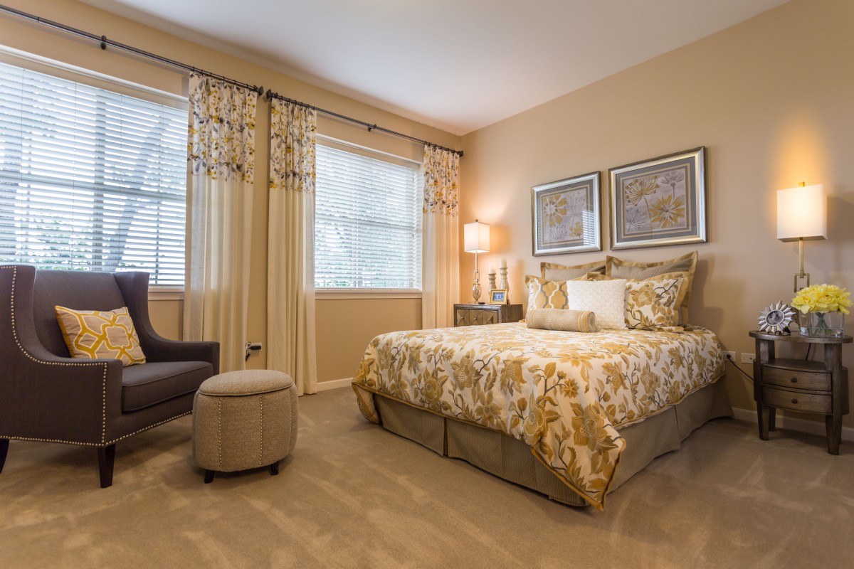A well-decorated bedroom featuring a bed with patterned bedding, matching curtains, a lamp, and a cozy chair.