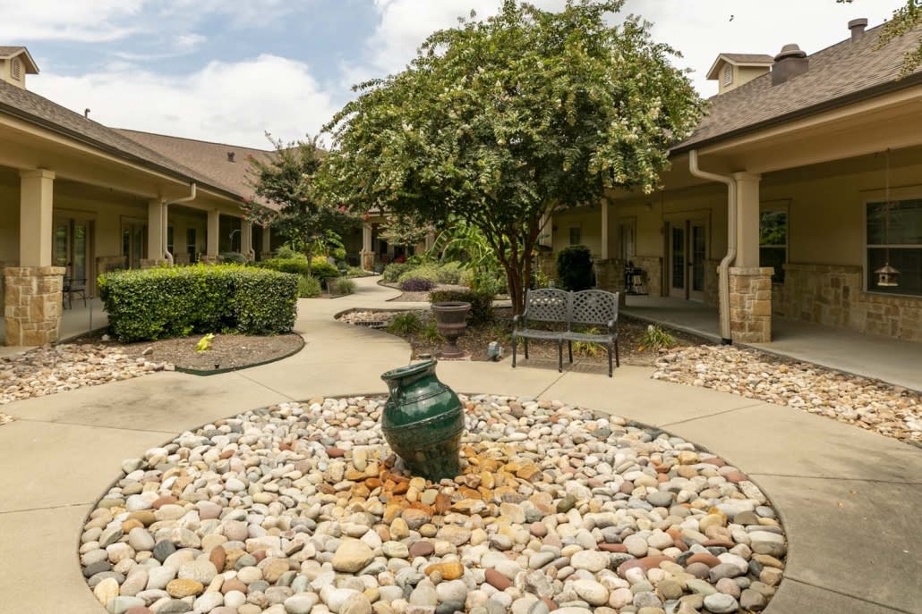 A serene courtyard with a central decorative vase surrounded by stones, lush greenery, and outdoor seating.