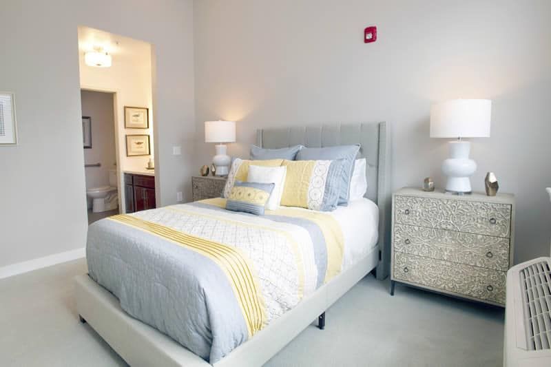 A cozy and well-decorated bedroom featuring a neatly made bed with gray and yellow bedding, flanked by matching nightstands and lamps.