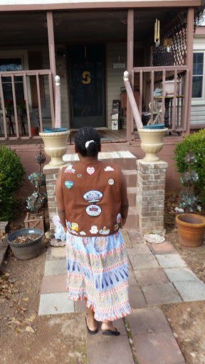 A person in a decorative vest and colorful skirt stands at the steps of a house, showcasing their unique style.