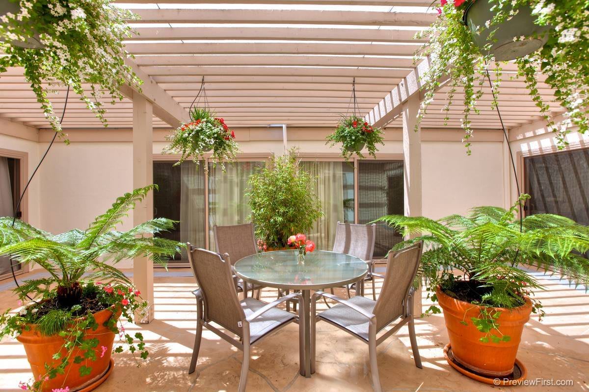 A cozy patio with a pergola, featuring a glass table and chairs, surrounded by lush greenery and hanging plants.
