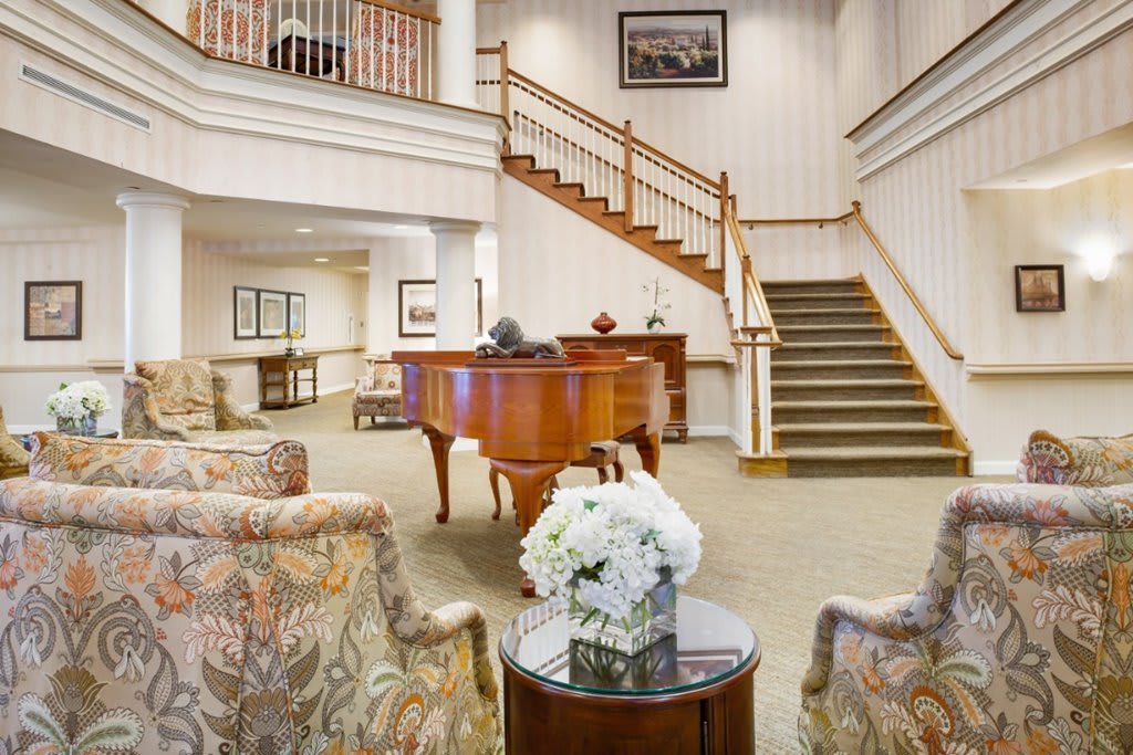 A spacious lobby featuring a grand piano, elegant staircase, and comfortable seating with floral decor.