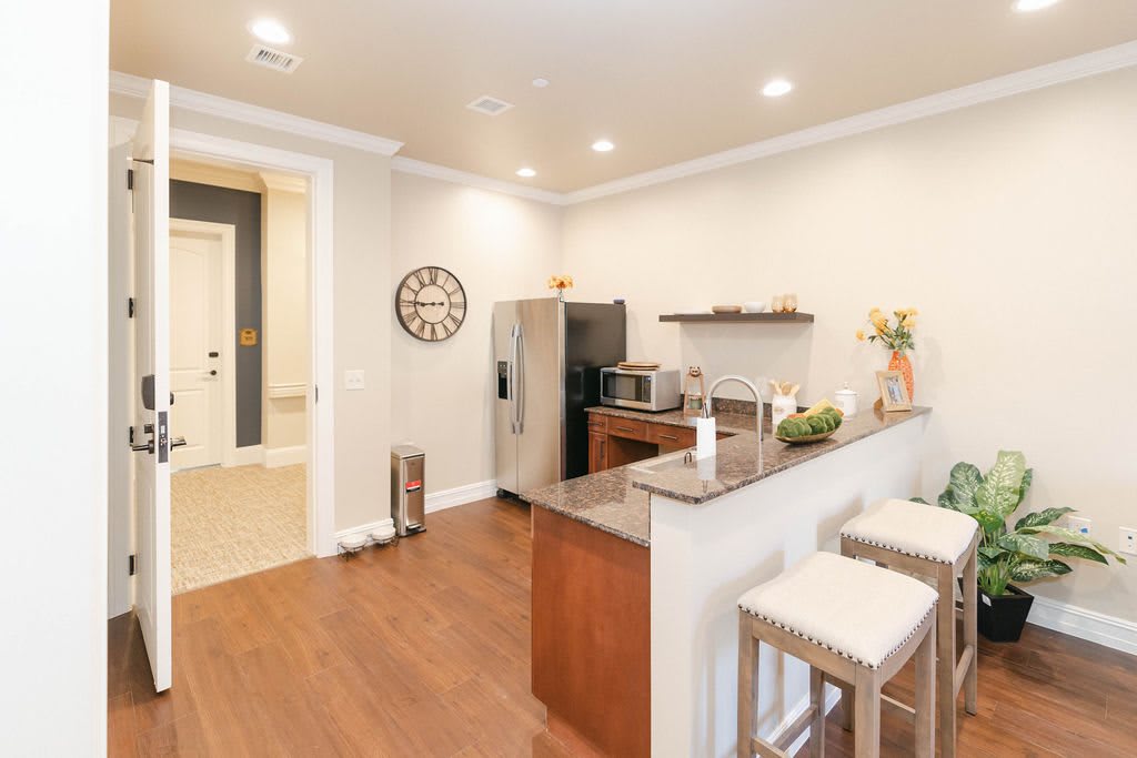 A modern kitchen interior with a minimalist design, featuring wood flooring, bar stools, and sleek appliances.