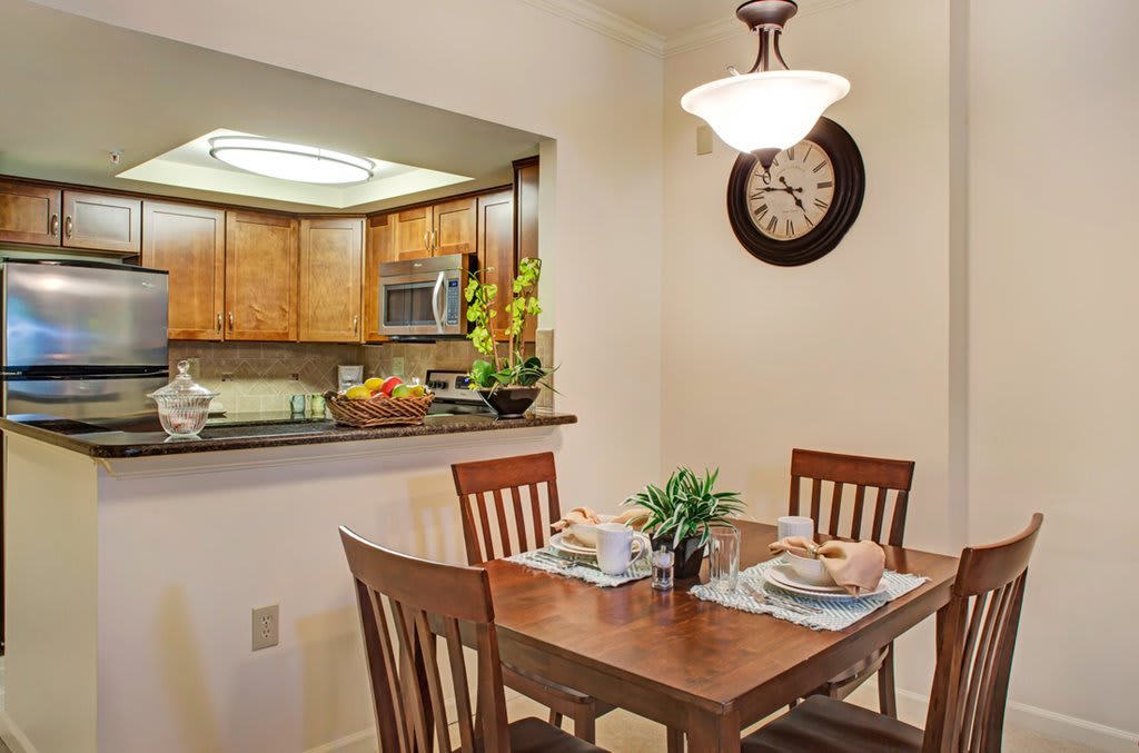 A cozy dining area with a wooden table and chairs, adjacent to a kitchen, featuring decorative plants and a wall clock.