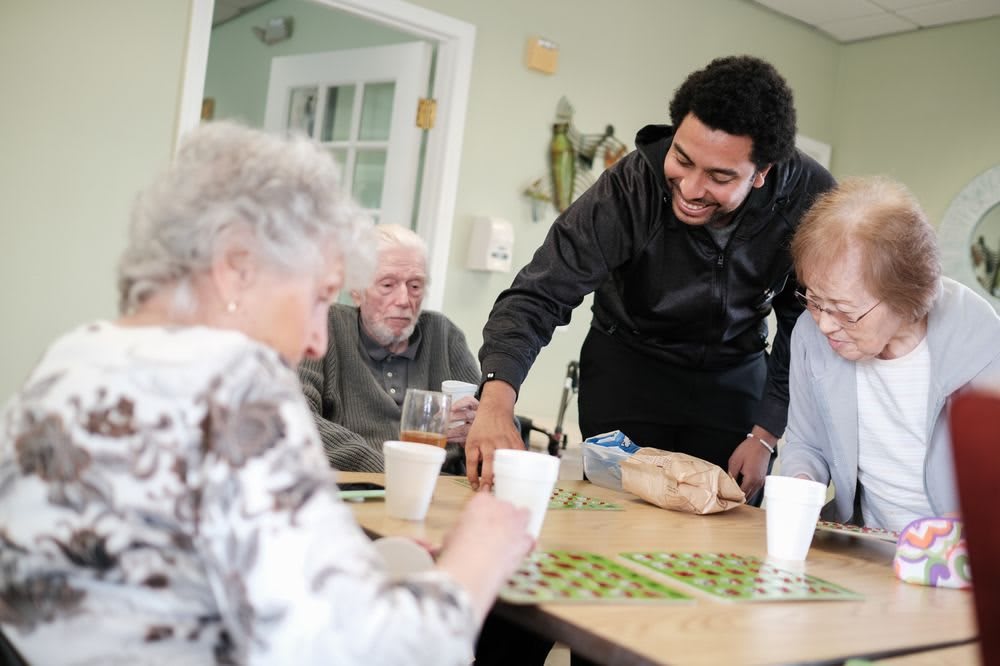 A caregiver interacts with elderly residents at a communal table.