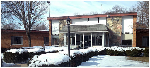 A snow-covered building in a winter landscape, featuring a lamp post and surrounding greenery.