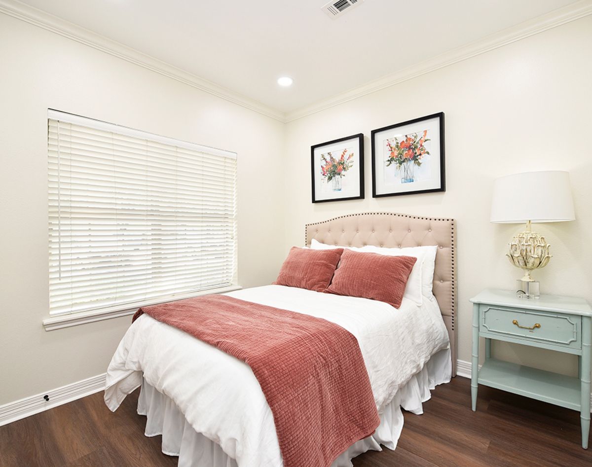 A cozy bedroom with a neatly made bed, accented with red pillows and a matching throw blanket. The room features two framed artworks above the headboard and a small nightstand with a lamp.