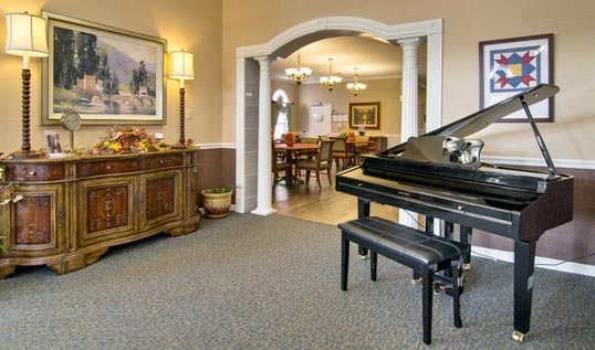 A cozy living room featuring a grand piano with a bench, elegantly decorated with artwork, furniture, and an archway leading to another room.