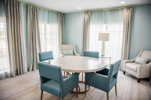 A modern dining area featuring a circular table surrounded by stylish blue chairs, illuminated by natural light filtering through sheer curtains.