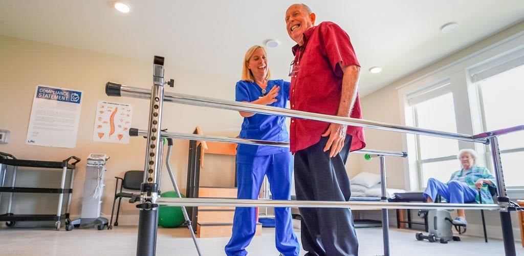 An elderly man is guided by a healthcare professional during a rehabilitation session using exercise bars.