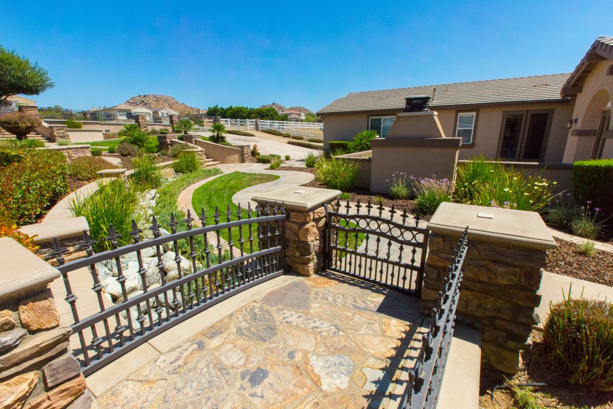 A suburban backyard with a stone-paved path, garden, and metal fencing under a clear blue sky.