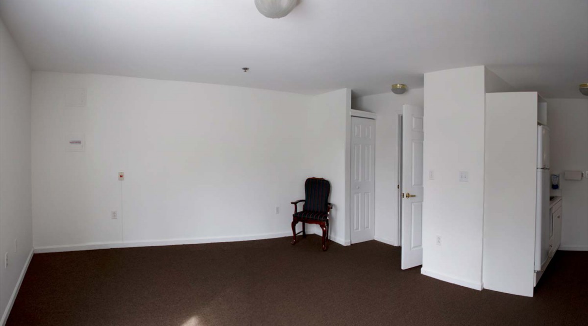 An empty room with white walls, a brown carpet, and a single chair near an open door.
