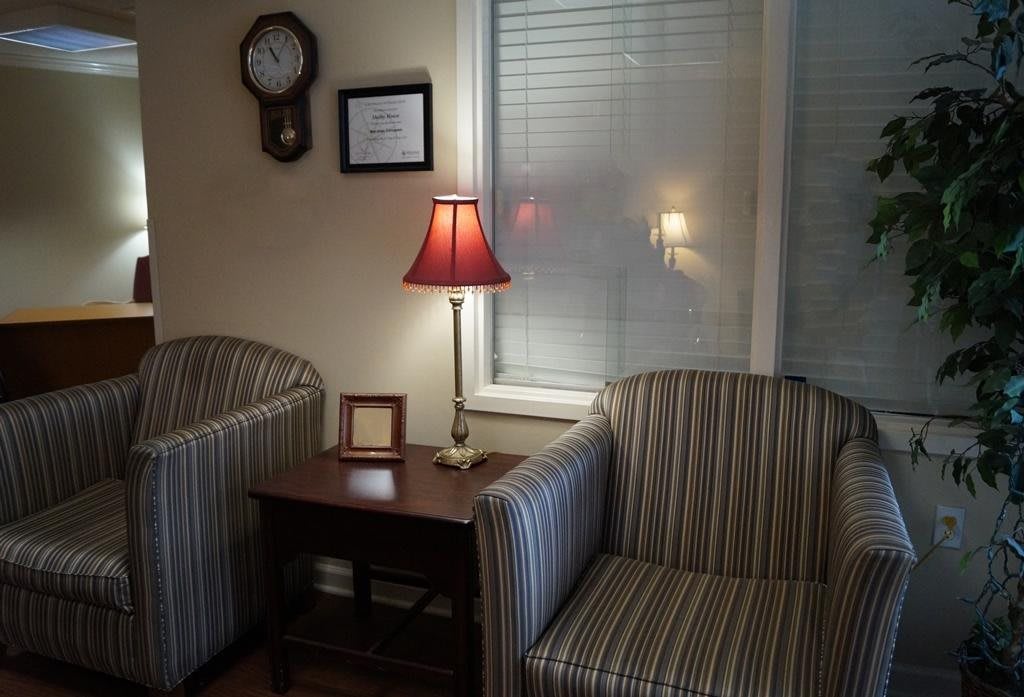 A cozy living room corner featuring two striped chairs, a side table with a red lampshade lamp, and a wall clock.