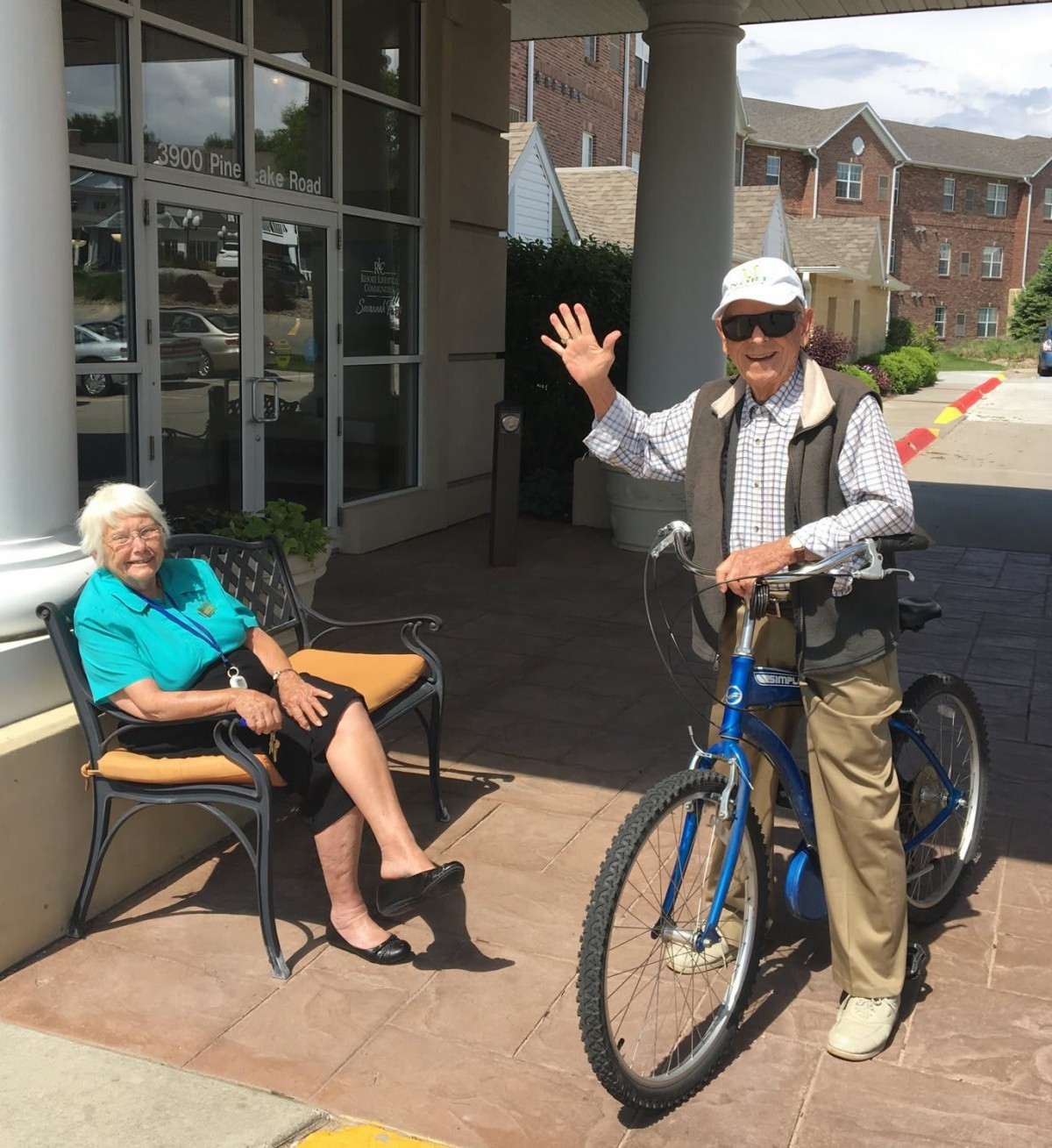 An elderly man with a bicycle waves while an elderly woman sits on a chair outside.
