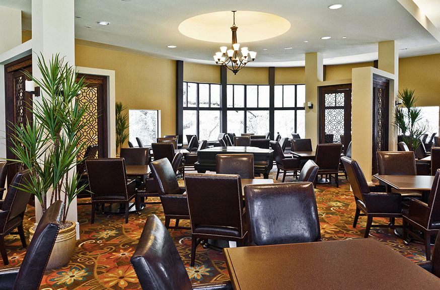 A well-furnished dining area with multiple tables and chairs, illuminated by a chandelier, featuring large windows and a colorful carpet.
