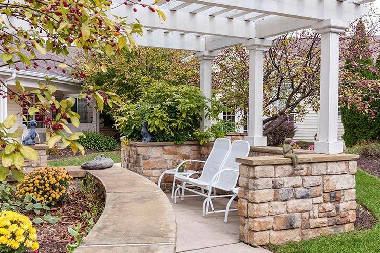 A cozy patio with a white pergola and chairs surrounded by lush greenery and stone walls.