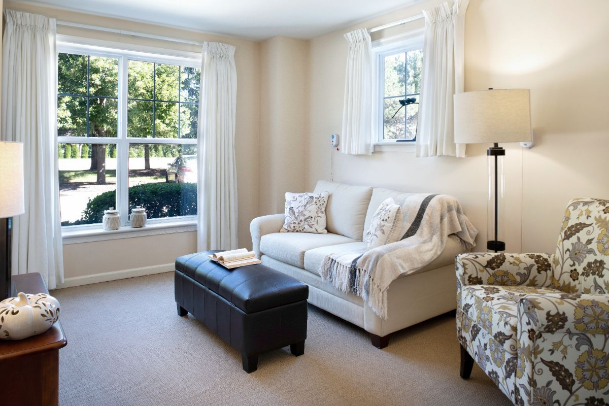 A cozy living room with a sofa, patterned chairs, and an ottoman by a large window letting in natural light.