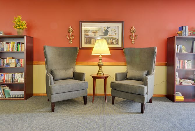 A cozy living room setup featuring two grey armchairs, a round wooden table with a lamp, and wall decor.