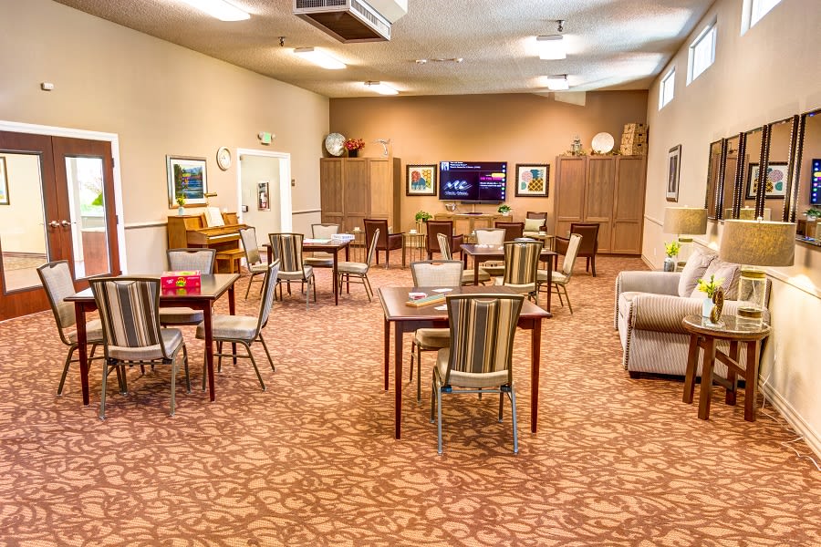 A well-lit community dining area with multiple tables and chairs, featuring a television on the wall.