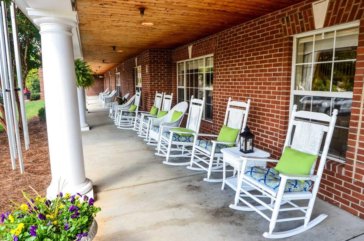 A charming porch with white rocking chairs and green cushions lined up along a brick wall, complemented by hanging plants.