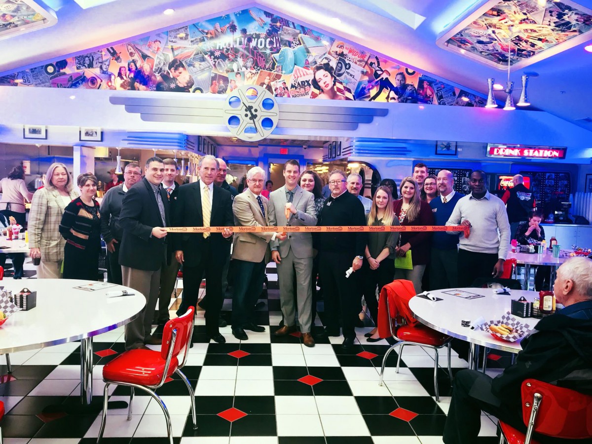 A group of people participate in a ribbon-cutting ceremony inside a vibrant, retro-themed diner.