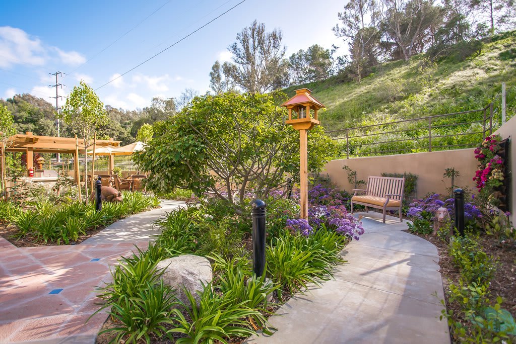 A scenic garden pathway lined with lush greenery, trees, and a wooden birdhouse, offering a serene outdoor space.