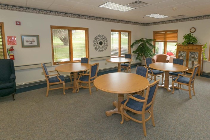 A bright and well-lit interior with several round tables and chairs arranged in a spacious manner, featuring large windows and a decorative plant.