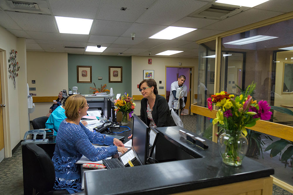 A busy hospital reception area with staff and visitors interacting at the front desk.