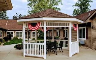 A decorated gazebo with outdoor furniture surrounded by trees and a building.