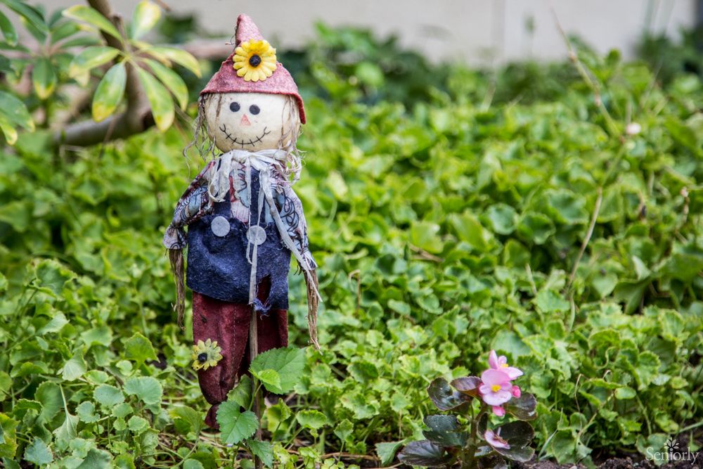 A small scarecrow stands in a lush garden surrounded by greenery and flowers.
