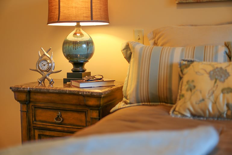 A cozy bedroom scene featuring a wooden nightstand adorned with a stylish lamp and decorative items, alongside a bed with striped pillows.