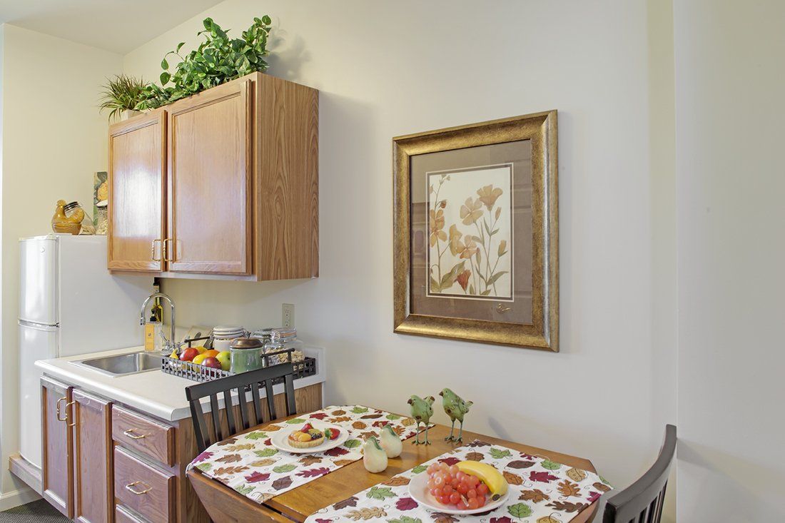A cozy kitchen corner with a small dining table set for two, adorned with a colorful tablecloth and a plate of fruit. A framed artwork hangs on the wall beside wooden cabinets.
