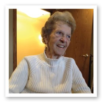 An elderly woman is sitting indoors, smiling warmly while wearing a white sweater.