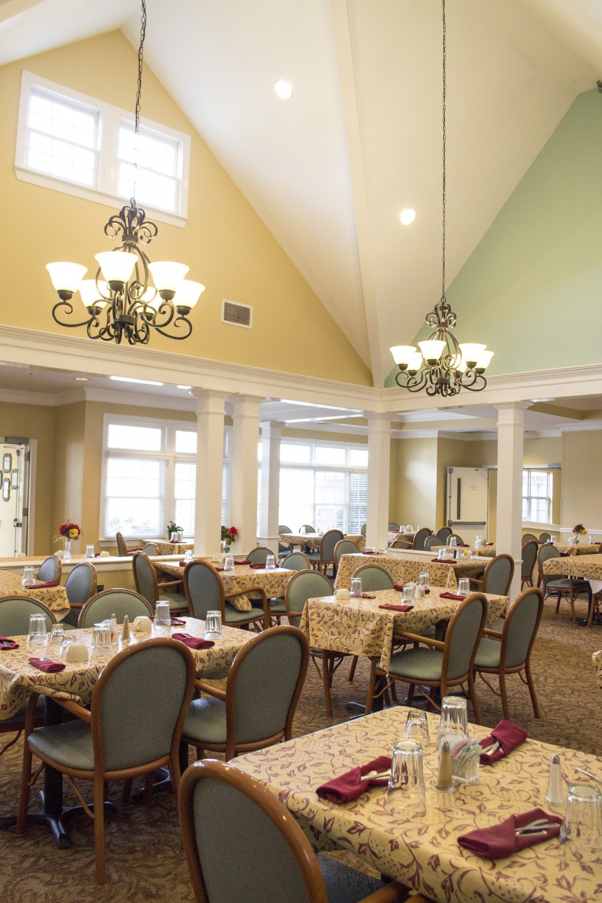 A spacious dining area with neatly arranged tables and chairs, illuminated by elegant chandeliers and natural light through large windows.
