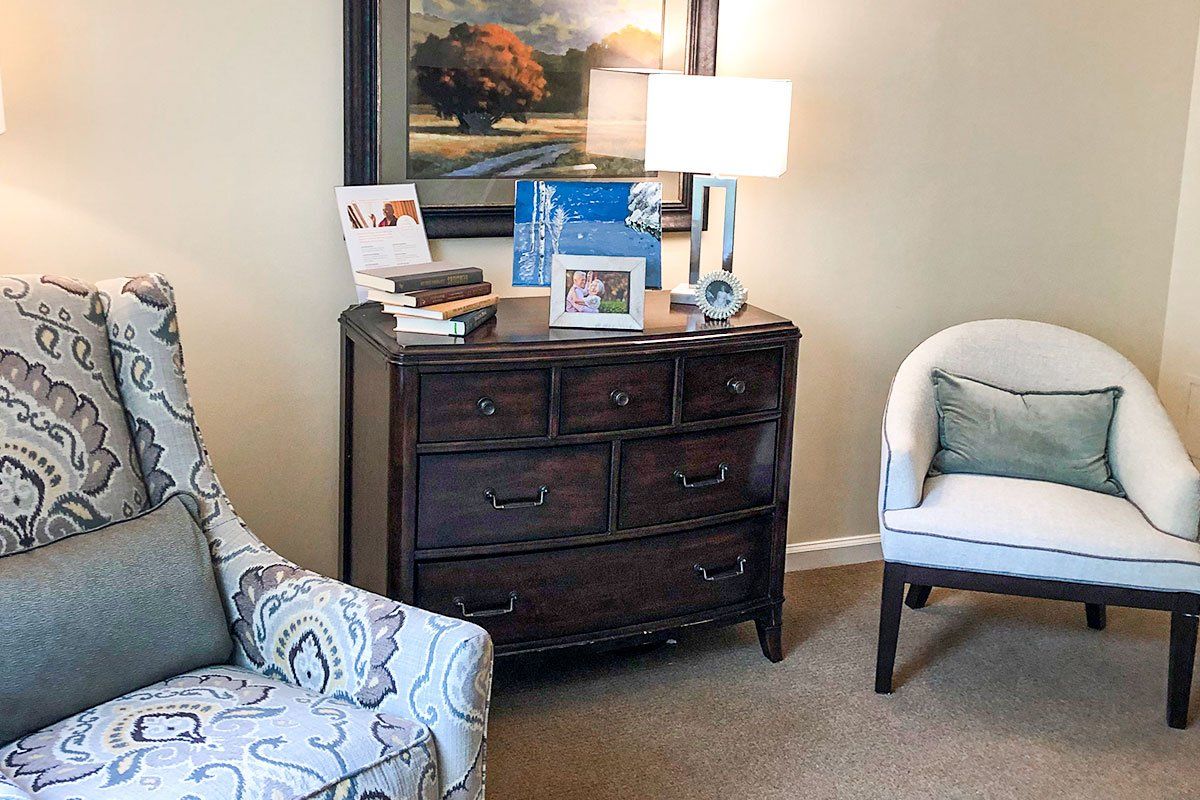 A cozy living room corner with a dark wooden dresser, a lamp on top, and a patterned armchair.