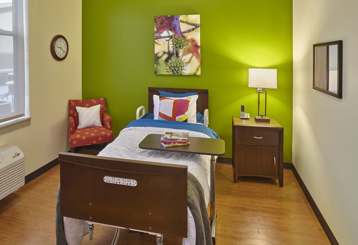 A cozy bedroom with a green accent wall, featuring a single bed, colorful artwork, and a warm lamp on the nightstand.
