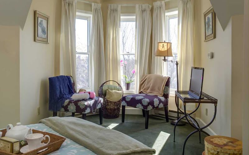 A cozy living room with large windows, two chairs, a floor lamp, and soft curtains.