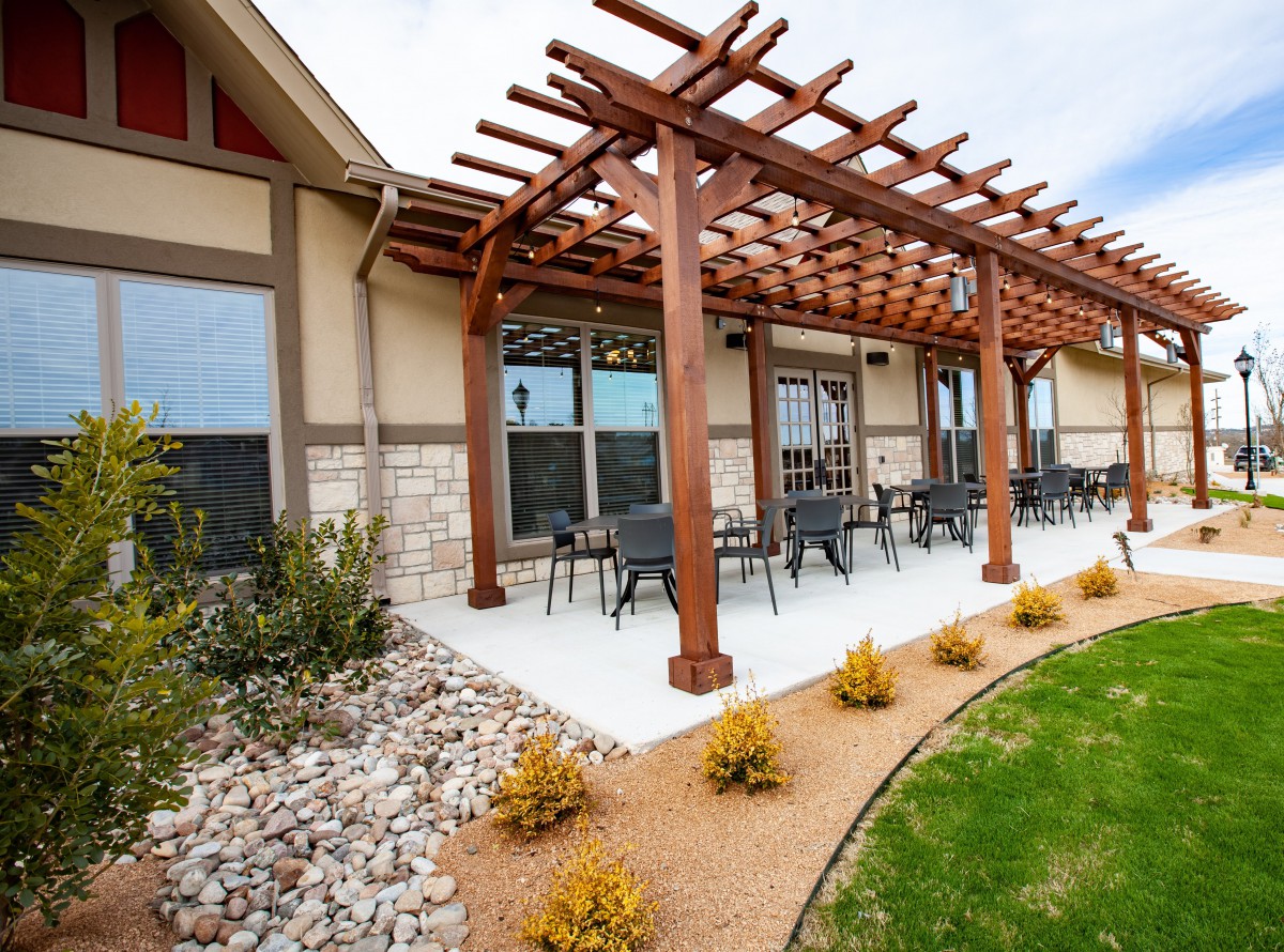 A modern patio with a wooden pergola and outdoor seating, surrounded by landscaped greenery.