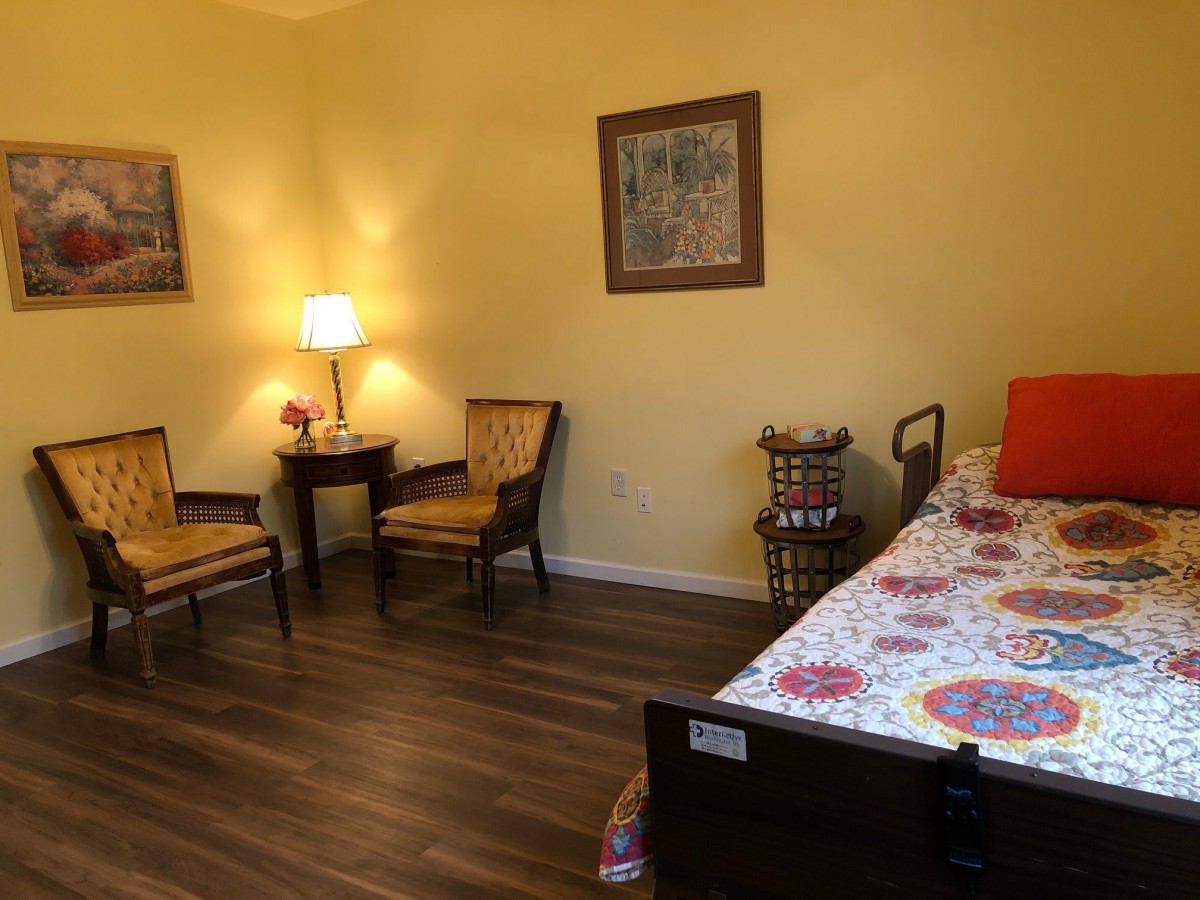A cozy bedroom with wooden flooring, featuring a bed, two chairs, a table with a lamp, and colorful decor.
