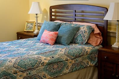 A cozy bedroom scene with a neatly made bed featuring patterned bedding, multiple pillows, a nightstand with a lamp, and a picture frame.