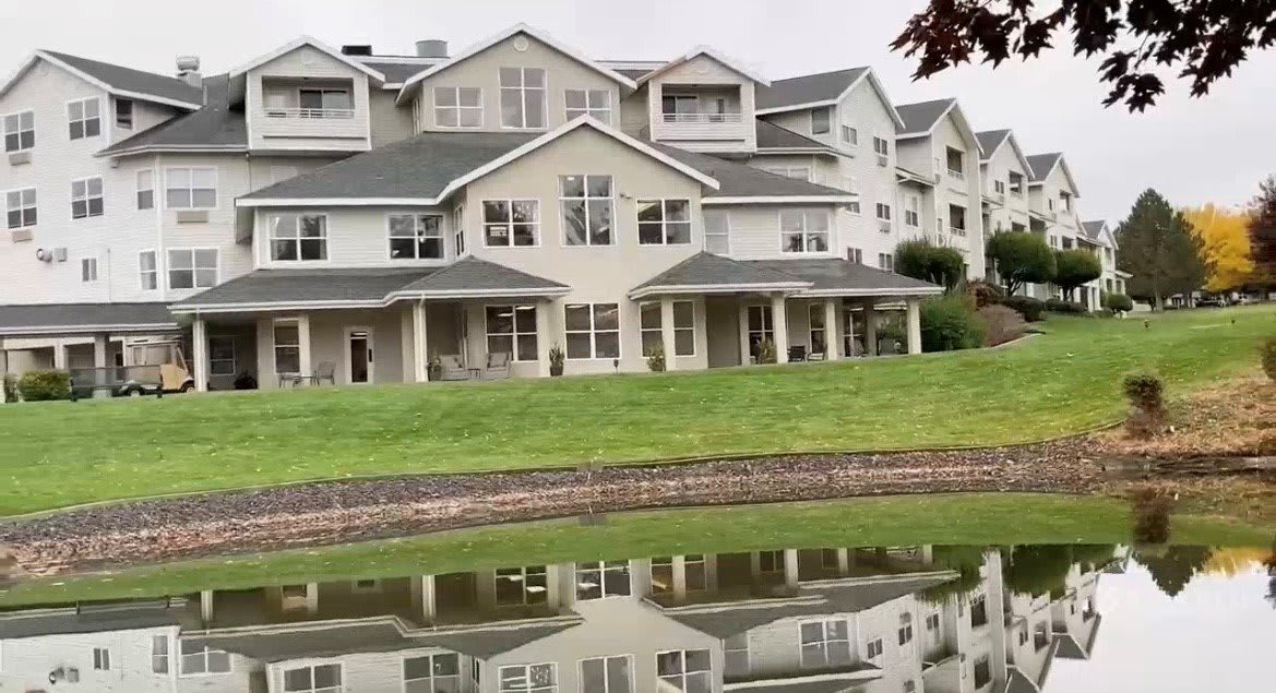 A large residential building with multiple stories is reflected in a pond, surrounded by a well-maintained lawn.