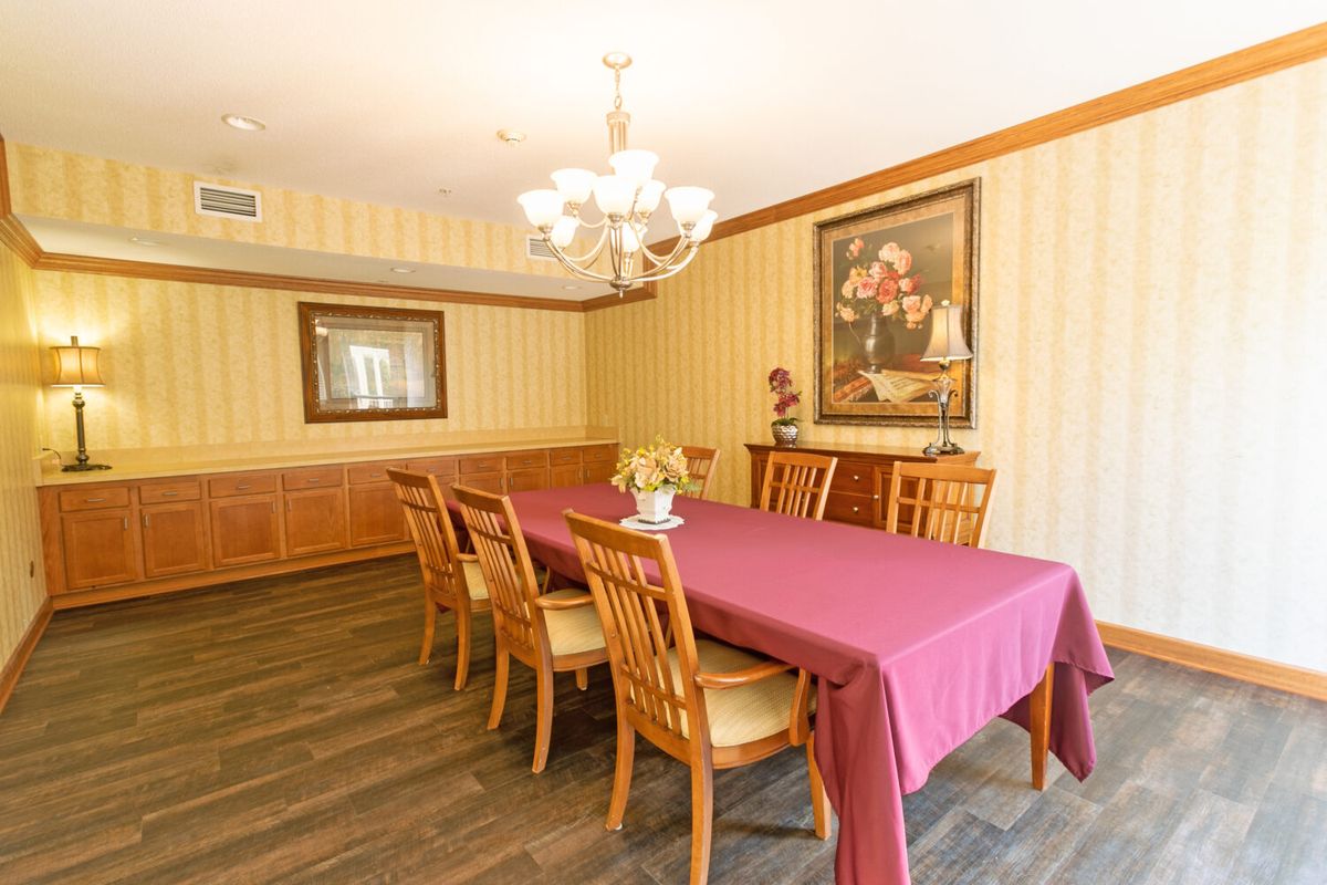 A dining room with a long table covered in a red tablecloth, surrounded by wooden chairs and adorned with a chandelier and artwork on the walls.