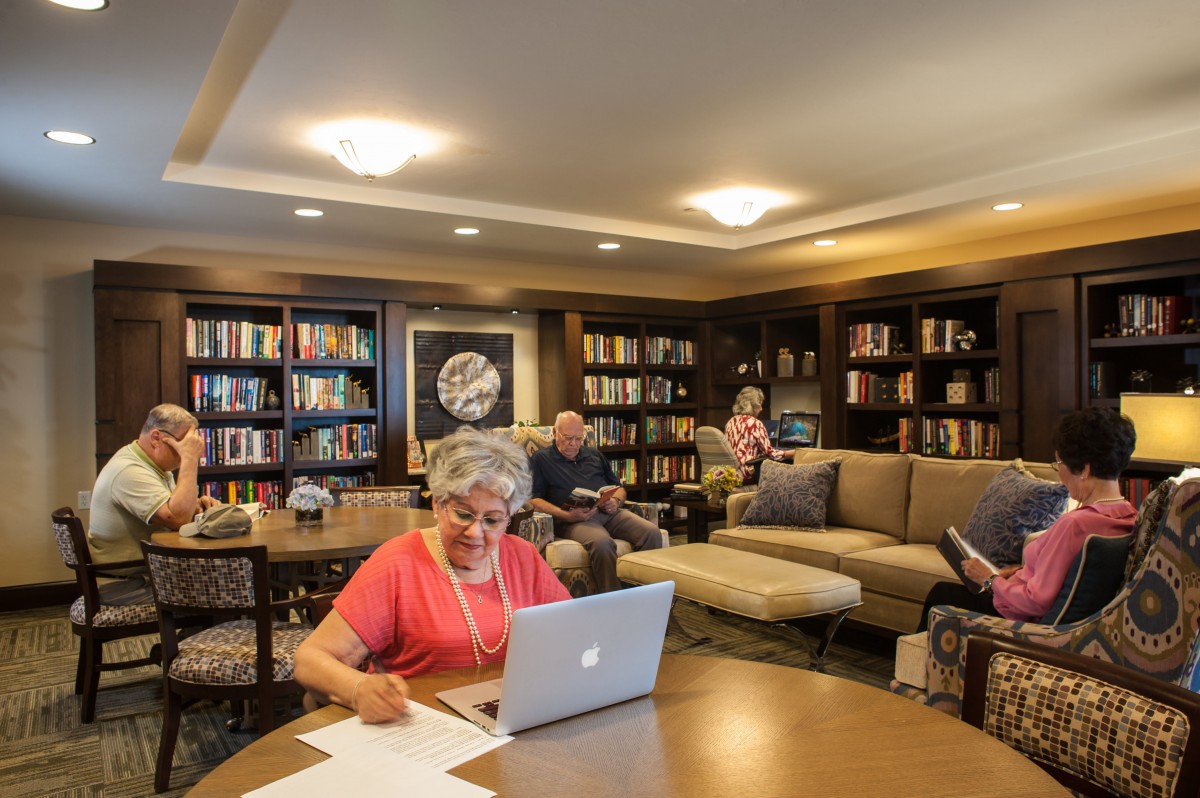 People reading and working in a cozy library setting with bookshelves and comfortable seating.