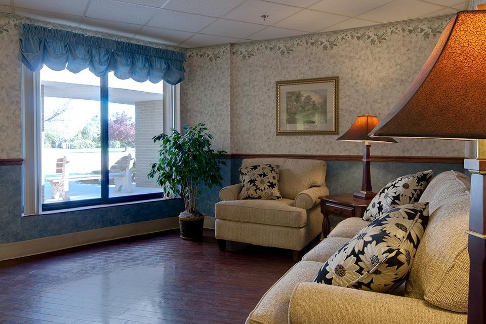 A cozy living room with a sofa, armchair, lamp, and large window overlooking an outdoor area.