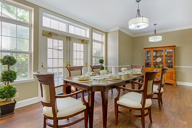 A well-lit dining room with a wooden table set and upholstered chairs, featuring large windows and elegant lighting.