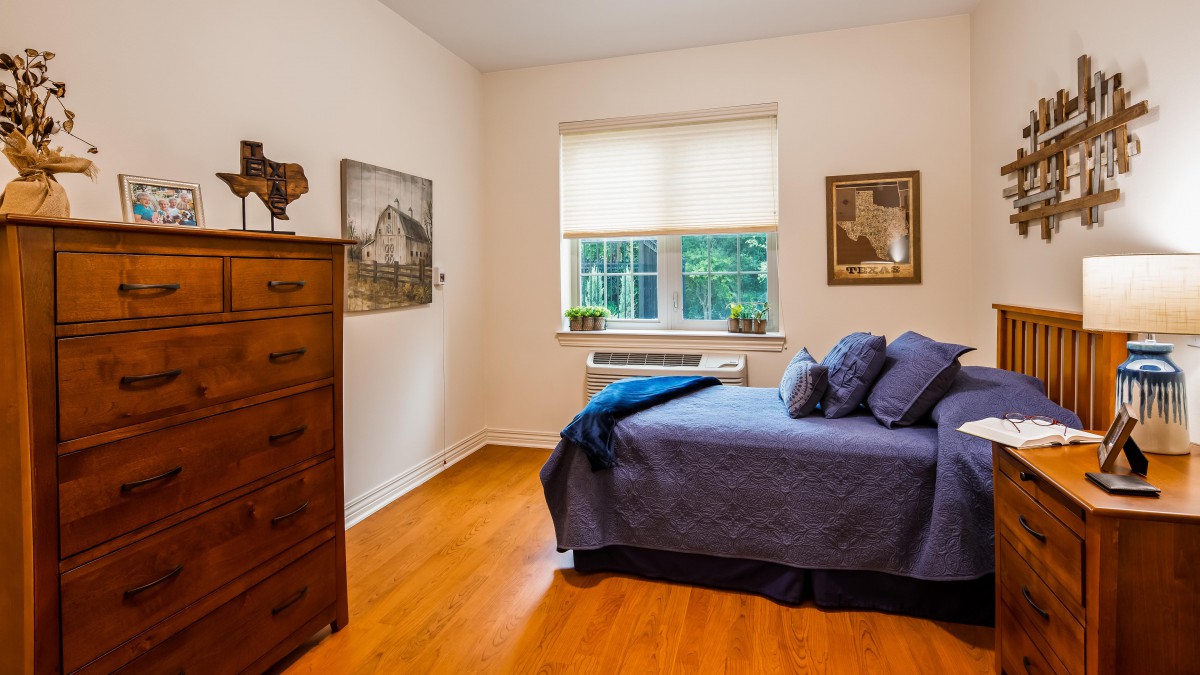 A cozy bedroom with a wooden dresser, a neatly made bed with pillows, and natural light streaming through the window.
