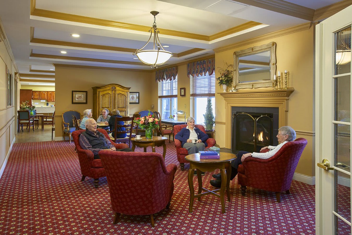A cozy lounge area with seniors seated around a fireplace.