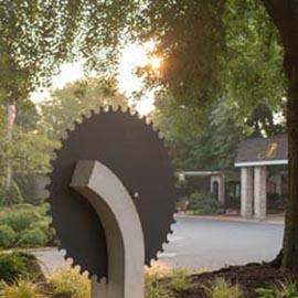 A modern sculpture resembling a saw blade, set against a lush garden backdrop with warm sunlight filtering through the trees.