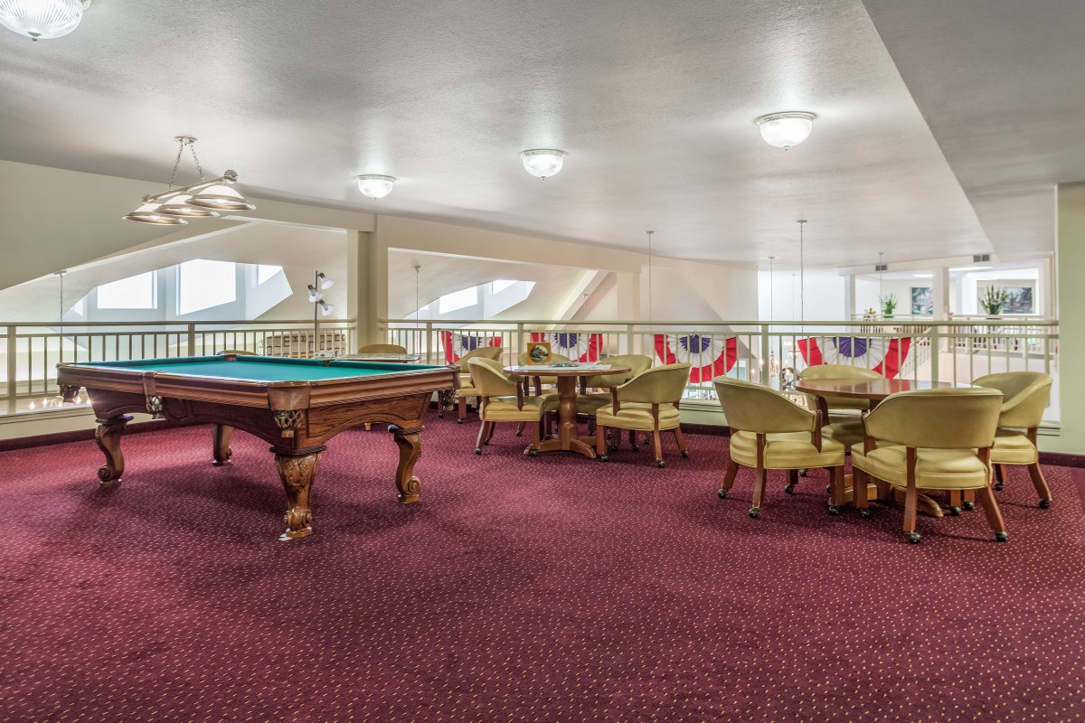 A spacious billiards room with a pool table and seating area, featuring red carpet and bright lighting.