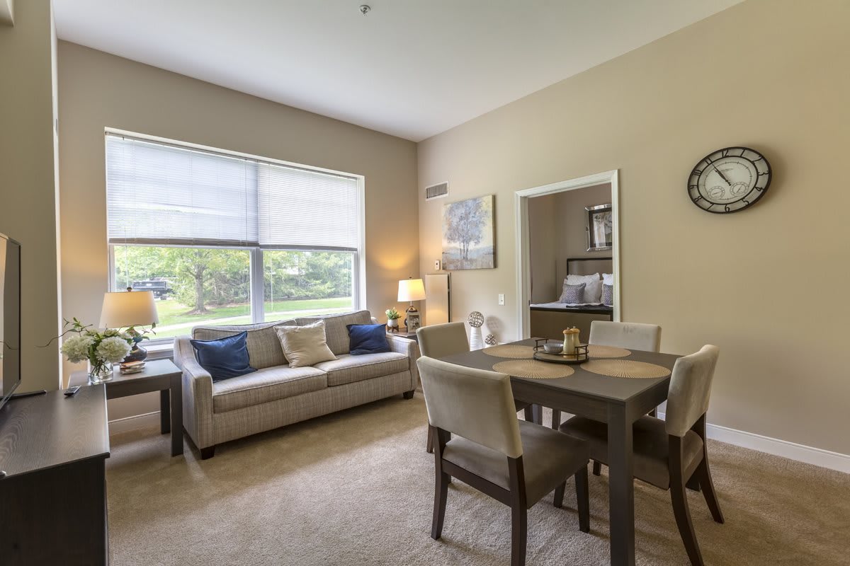 A cozy living room and dining area with a large window, featuring a comfortable sofa, dining table and chairs, and decorative elements including a lamp, artwork, and clock.